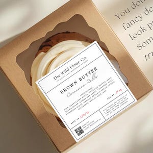 May include: A brown cardboard box with a clear window, containing a cinnamon roll. The label reads "Brown Butter Cinnamon Rolls" from The Wild Flour Co. Text on the box says, "You don't need a fancy design to look put together. Sometimes less truly is more."
