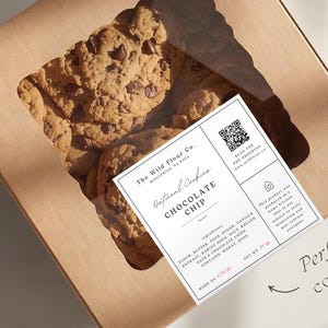 Puede incluir: Una caja de cartón marrón con una ventana transparente, llena de galletas con chispas de chocolate. Una etiqueta blanca con texto negro está en la caja, que enumera los ingredientes y un código QR. El texto de la etiqueta dice "Oatmeal Cookies Chocolate Chip".