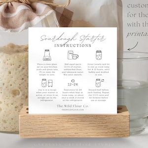 May include: A clear glass jar with a beige and white checkered cloth tied around the top, containing a sourdough starter. A card with sourdough instructions sits in a wooden holder, next to a glass measuring cup.