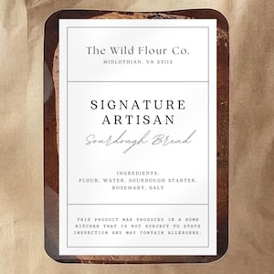 Könnte beinhalten: Eine braune Papiertüte mit einem weißen Etikett mit der Aufschrift "The Wild Flour Co." und "Signature Artisan Sourdough Bread". Zutaten: Mehl, Wasser, Sauerteig, Rosmarin und Salz.