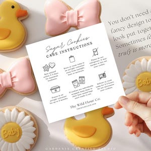 May include: A square white card with black text titled Sugar Cookies CARE INSTRUCTIONS, surrounded by decorated cookies shaped like yellow ducks, pink bows, and white daisies with the word Baby in the center, all on a light surface.