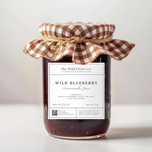 Op de afbeelding: Een glazen pot gevuld met donkerpaarse, zelfgemaakte bosbessenjam. Het deksel is bedekt met een bruin-witte gingham stoffen hoes, vastgebonden met touw. Het etiket vermeldt The Wild Flour Co. Wild Blueberry Homemade Jam.