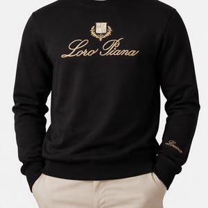 K&ouml;nnte beinhalten: Schwarzes Sweatshirt mit Rundhalsausschnitt und goldfarbener &bdquo;Loro Piana&ldquo;-Stickerei und Emblem auf der Brust. Das lang&auml;rmlige Oberteil hat einen gerippten Saum und B&uuml;ndchen, und der Markenname ist auch auf dem linken &Auml;rmel gestickt.