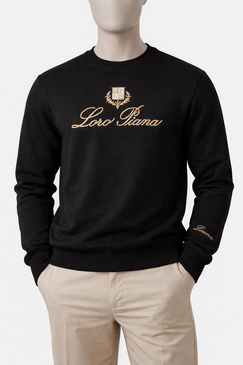 K&ouml;nnte beinhalten: Schwarzes Sweatshirt mit Rundhalsausschnitt und goldfarbener &bdquo;Loro Piana&ldquo;-Stickerei und Emblem auf der Brust. Das lang&auml;rmlige Oberteil hat einen gerippten Saum und B&uuml;ndchen, und der Markenname ist auch auf dem linken &Auml;rmel gestickt.