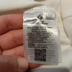 K&ouml;nnte beinhalten: Nahaufnahme eines wei&szlig;en Stoffetiketts mit dem Markennamen "Loro Piana" und mehrsprachigem Text. Das Etikett enth&auml;lt einen QR-Code und Informationen &uuml;ber die Zusammensetzung und Herkunft des Stoffes.
