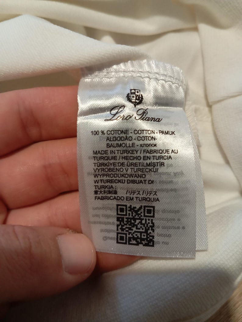 K&ouml;nnte beinhalten: Nahaufnahme eines wei&szlig;en Stoffetiketts mit dem Markennamen "Loro Piana" und mehrsprachigem Text. Das Etikett enth&auml;lt einen QR-Code und Informationen &uuml;ber die Zusammensetzung und Herkunft des Stoffes.