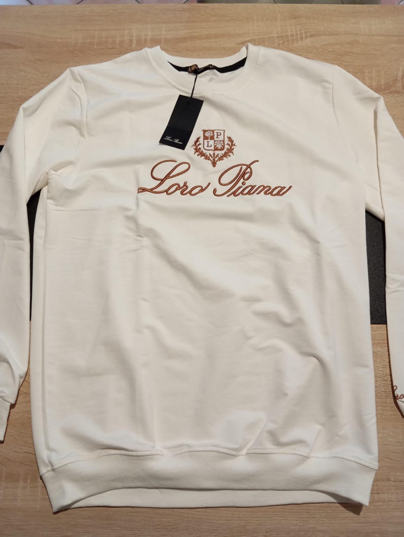 K&ouml;nnte beinhalten: Wei&szlig;es Sweatshirt mit langen &Auml;rmeln. Auf der Vorderseite ist der Markenname "Loro Piana" in brauner Schrift gestickt, zusammen mit einem Wappen. Ein schwarzes Etikett h&auml;ngt am Ausschnitt. Das Sweatshirt ist aus weichem Material.