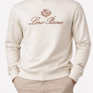 Peut inclure: Sweat-shirt crème avec le texte brodé "Loro Piana" en script marron. Le sweat-shirt présente un petit emblème au-dessus du texte et le nom de la marque sur le poignet. Le modèle porte un pantalon kaki.