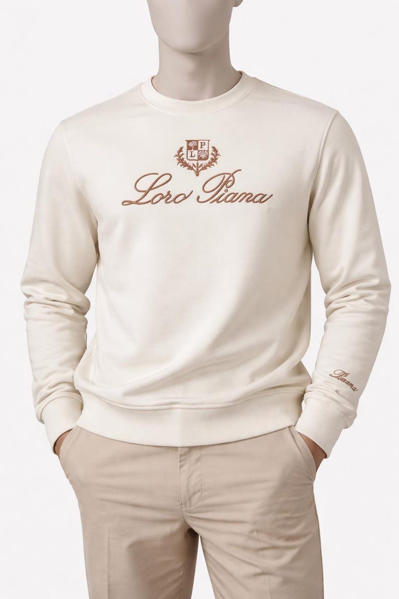 K&ouml;nnte beinhalten: Cremefarbenes Sweatshirt mit dem gestickten Text "Loro Piana" in brauner Schrift. Das Sweatshirt hat ein kleines Emblem &uuml;ber dem Text und den Markennamen am &Auml;rmel. Das Model tr&auml;gt eine khakifarbene Hose.