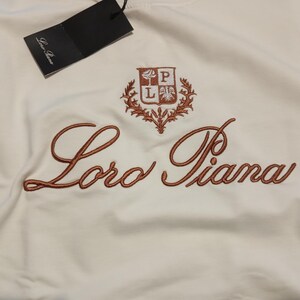 K&ouml;nnte beinhalten: Wei&szlig;es Kleidungsst&uuml;ck mit gesticktem Schriftzug "Loro Piana" in warmem Braun. Ein passendes Emblem befindet sich &uuml;ber der Schrift. Ein schwarzes Etikett h&auml;ngt oben.