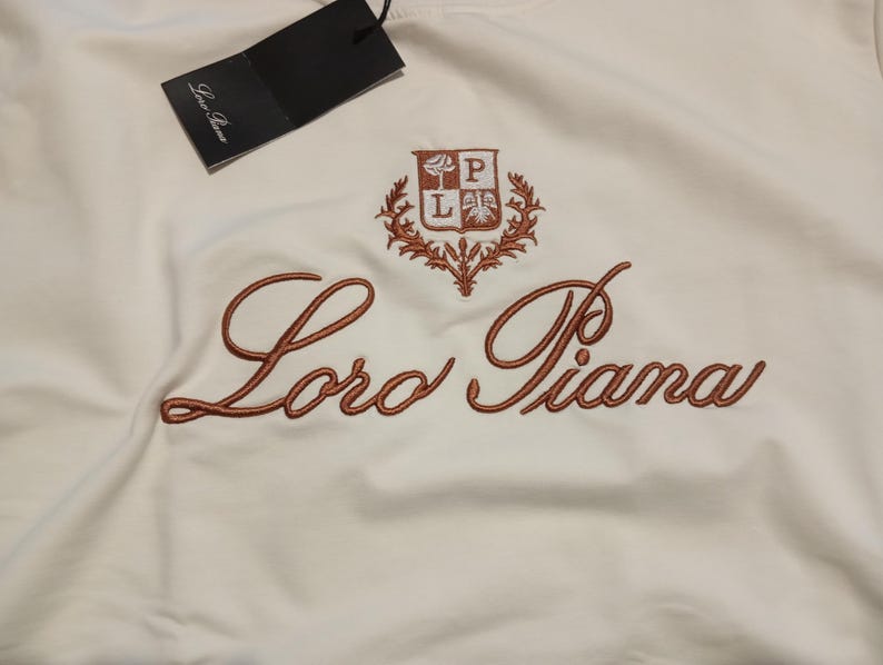 K&ouml;nnte beinhalten: Wei&szlig;es Kleidungsst&uuml;ck mit gesticktem Schriftzug "Loro Piana" in warmem Braun. Ein passendes Emblem befindet sich &uuml;ber der Schrift. Ein schwarzes Etikett h&auml;ngt oben.