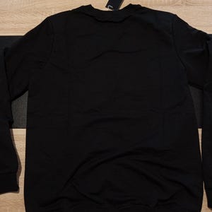 K&ouml;nnte beinhalten: Ein schwarzes Sweatshirt mit Rundhalsausschnitt und langen &Auml;rmeln. Ein schwarzes Etikett ist am Kragen befestigt. Das Sweatshirt ist aus einem weichen Material gefertigt.