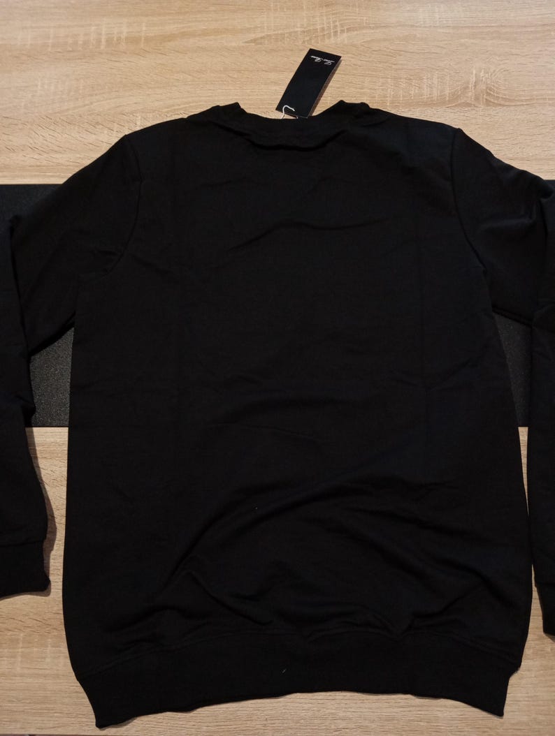 K&ouml;nnte beinhalten: Ein schwarzes Sweatshirt mit Rundhalsausschnitt und langen &Auml;rmeln. Ein schwarzes Etikett ist am Kragen befestigt. Das Sweatshirt ist aus einem weichen Material gefertigt.