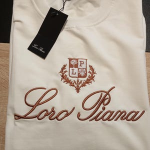 K&ouml;nnte beinhalten: Cremefarbenes T-Shirt mit dem gestickten Markennamen "Loro Piana" in Schreibschrift. Das Shirt hat ein kleines Emblem &uuml;ber dem Markennamen. Ein schwarzes Etikett h&auml;ngt am Kragen.