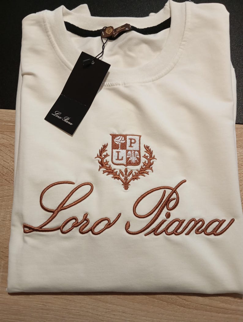 K&ouml;nnte beinhalten: Cremefarbenes T-Shirt mit dem gestickten Markennamen "Loro Piana" in Schreibschrift. Das Shirt hat ein kleines Emblem &uuml;ber dem Markennamen. Ein schwarzes Etikett h&auml;ngt am Kragen.