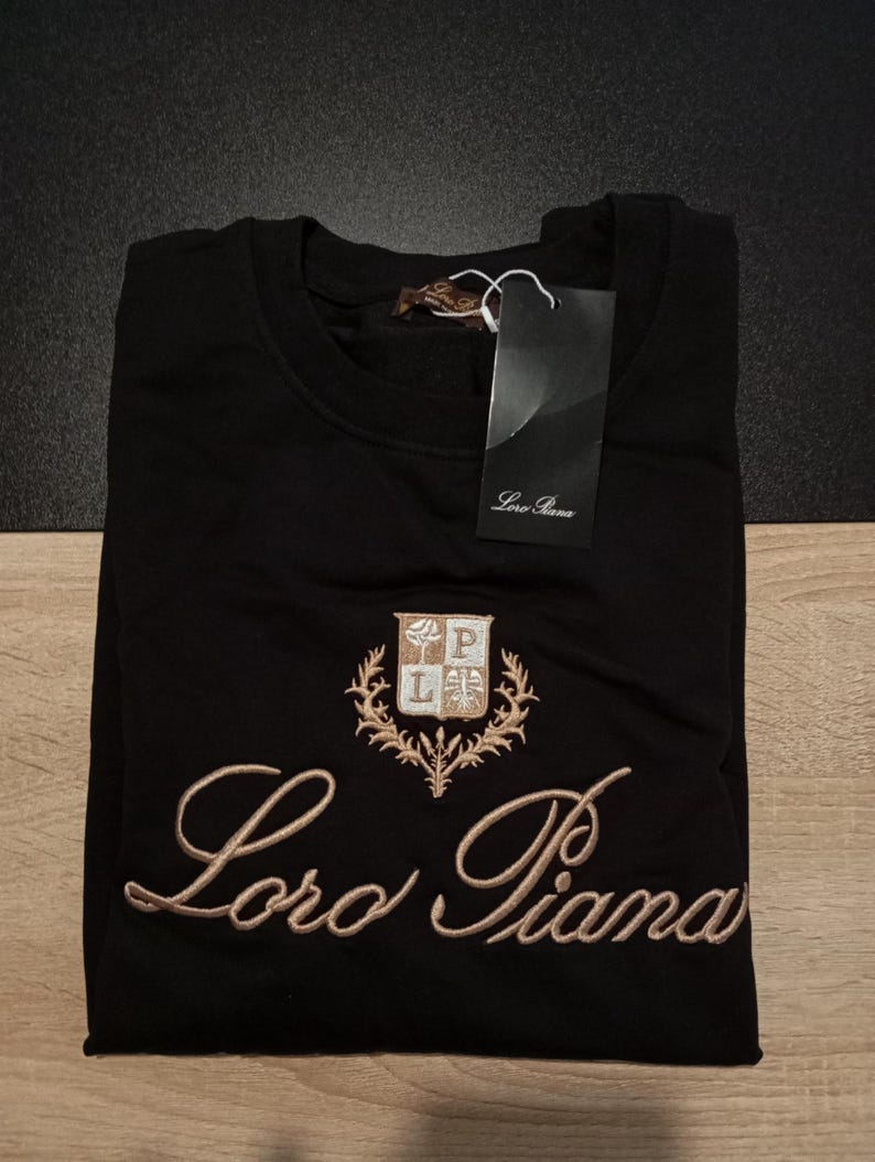 K&ouml;nnte beinhalten: Schwarzes Sweatshirt mit gesticktem "Loro Piana"-Schriftzug und Wappenlogo. Das Wappen zeigt die Buchstaben "L" und "P" mit einem dekorativen Rand. Ein Etikett mit dem Markennamen ist angebracht.