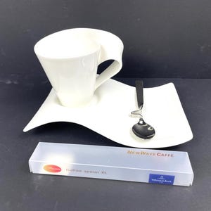May include: A white ceramic coffee cup and saucer set with a unique wave design. A stainless steel spoon rests on the saucer. A box of coffee spoons is in the foreground. The set is from the NewWave Caffe collection.