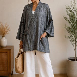 May include: A woman wearing a gray kimono-style jacket with a vertical striped pattern, white pants, and a straw tote bag. The jacket has pockets and a tie closure. The outfit is paired with brown sandals.
