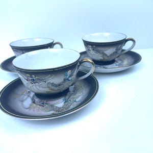 May include: Set of three Japanese tea cups and saucers. The cups and saucers are black with a dragon design in shades of gray, blue, and gold. The cups have gold handles and a white interior. The saucers have a matching design.