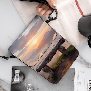 May include: A rectangular crossbody bag with a sunset landscape design. The bag features a black strap and a flap closure. The image on the bag depicts a sunset over a wooden fence. A black mug and a magazine are also visible.