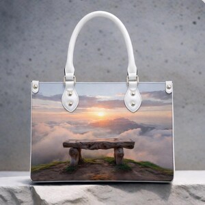 May include: Tote bag with a scenic landscape print. The image features a wooden bench on a hilltop overlooking a sunset above the clouds. The bag has white handles and silver-tone hardware.