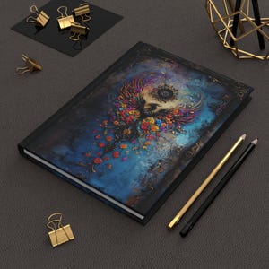 May include: A black journal with a colorful phoenix design on the cover. The phoenix is surrounded by flowers and has a large eye in the center of its chest. The journal has a black spine and a gold-colored trim.