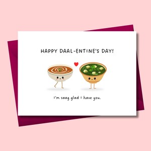 Könnte beinhalten: Eine Valentinstagskarte mit dem Text "HAPPY DAAL-ENTINE'S DAY!" und dem Satz "I'm saag glad I have you." Dargestellt sind zwei Cartoon-Schüsseln mit Essen, eine mit brauner Suppe und die andere mit grüner Suppe und Tofu.