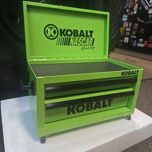 May include: A bright green Kobalt tool chest with two drawers and a top compartment. The Kobalt and NASCAR Racing logos are prominently displayed. The tool chest is on wheels and has a black handle.