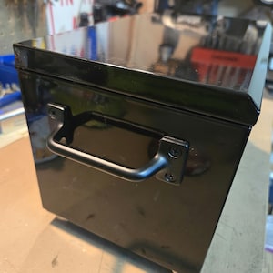 May include: Black metal toolbox with a handle and wheels. The toolbox has a shiny finish.