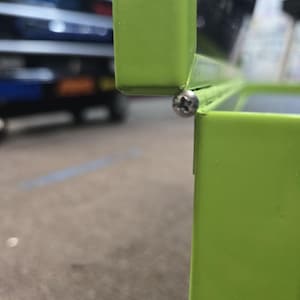 May include: Close-up of a bright green metal object, possibly a container or part of a larger structure. A silver screw fastens two sections together. The background is blurred, suggesting an outdoor setting.