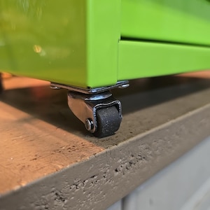 May include: Close-up of a green metal cabinet with a black caster wheel. The cabinet has a silver metal frame and a black rubber wheel.