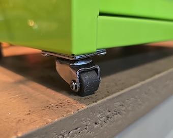 May include: Close-up of a green metal cabinet with a black caster wheel. The cabinet has a silver metal frame and a black rubber wheel.
