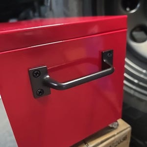 May include: A red metal toolbox with a black handle and wheels. The toolbox is sitting on a cardboard box with the word "KOMAX" printed on it.