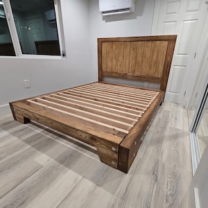 May include: A wooden bed frame with a headboard. The bed frame is made of stained wood and features a slatted base. The headboard is constructed from vertical wooden planks. The bed frame is set on a light-colored wood floor.