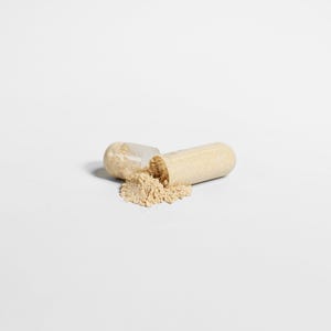 May include: An open, beige capsule with a light brown powder spilling out onto a white surface. The capsule is split in two, with one half lying next to the other. The powder is a fine, granular substance.