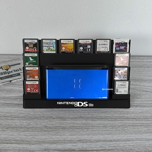 Könnte beinhalten: Ein schwarzer Kunststoff-Präsentationsständer für eine Nintendo DS Lite-Konsole und zwölf Spielkassetten. Der Ständer zeigt das Nintendo DS Lite-Logo in weißem Text auf der Vorderseite und hält die blaue Handheld-Konsole in der Mitte.