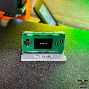 May include: A green handheld gaming console with a black screen displaying the text @my3Dprintz, a black directional pad, and black A and B buttons. The console rests on a grey stand with the embossed text GAME BOY ADVANCE on the front base.