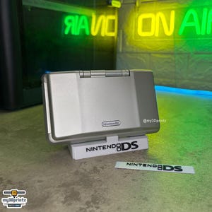 May include: A silver Nintendo DS handheld gaming console resting on a white plastic display stand. The stand features the black text NINTENDO DS, and a matching white rectangular plaque with the same text is placed in front of the console.