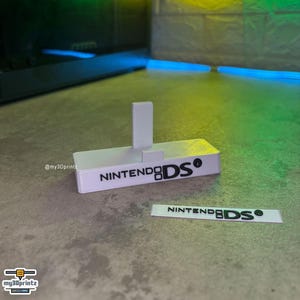 Könnte beinhalten: Ein weißer, rechteckiger Kunststoff-Präsentationsständer für eine Handheld-Spielkonsole mit einer vertikalen Stützlasche und dem schwarz aufgedruckten Text NINTENDO DS. Ein separater weißer, rechteckiger Streifen mit dem schwarzen Text NINTENDO DS liegt bei.