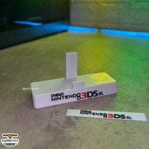 May include: A white 3D-printed display stand for a handheld gaming console, featuring a vertical support and a small front tab. The front of the stand displays the black and red text "new NINTENDO 3DS XL". A matching rectangular nameplate is included.