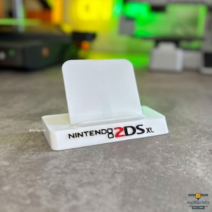 May include: A white plastic display stand featuring a rectangular base and an upright support panel. The front of the base displays the text NINTENDO 2DS XL in raised black lettering, with the number 2 highlighted in red.