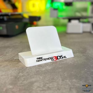 Peut inclure: Un support de présentation en plastique blanc pour une console de jeu portable. La base présente le texte noir et rouge "new NINTENDO 3DS XL" imprimé sur le devant. Le support possède un dossier rectangulaire et une base large.