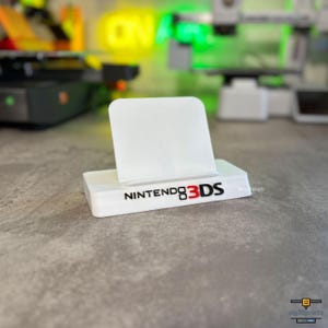 Könnte beinhalten: Ein weißer Kunststoff-Präsentationsständer für eine Nintendo 3DS Handheld-Konsole. Auf der Vorderseite der Basis ist das schwarze und rote Nintendo 3DS-Logo aufgedruckt. Der Ständer hat eine rechteckige Basis und eine geneigte Rückenstütze.