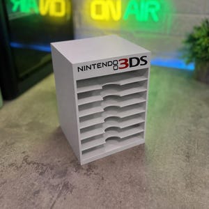 Könnte beinhalten: Ein weißer Aufbewahrungsturm aus Kunststoff für Nintendo 3DS-Spielhüllen mit acht horizontalen Fächern, die halbkreisförmige Aussparungen für den Zugriff bieten. An der oberen Vorderkante befindet sich das schwarz-rote Nintendo 3DS-Logo.