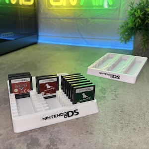 May include: A white plastic storage rack for Nintendo DS game cartridges featuring the Nintendo DS logo on the front. The rack holds several black game cartridges with labels including Cars and Nintendogs. A second empty rack is visible nearby.