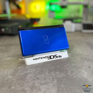 Könnte beinhalten: Eine kobaltblaue Nintendo DS Lite Handheld-Spielkonsole auf einem weißen Kunststoff-Präsentationsständer. Der Ständer trägt den schwarz aufgedruckten Text NINTENDO DS Lite. Die Konsole ist geschlossen und zeigt ihre glänzende Oberfläche.