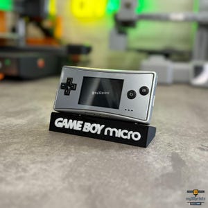 Könnte beinhalten: Eine silberne Game Boy Micro Handheld-Videospielkonsole auf einem schwarzen Ständer. Auf dem Ständer steht in weißer Schrift "GAME BOY micro". Die Konsole hat einen kleinen Bildschirm und schwarze Bedientasten. Das Bild enthält den Text "@my3Dprintz".