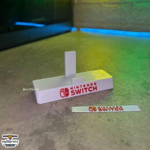 Könnte beinhalten: Ein weißer, 3D-gedruckter Nintendo Switch Konsolenständer mit einem roten Nintendo Switch Logo und Schriftzug auf der Vorderseite. Ein separates, dünnes weißes Rechteck mit demselben roten Logo und Schriftzug liegt vor dem Ständer.
