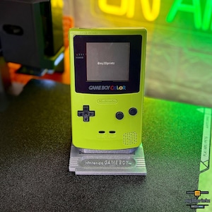 Könnte beinhalten: Eine limettengrüne Game Boy Color Handheld-Spielkonsole auf einem grauen, 3D-gedruckten Ständer in Form einer Spielkassette. Die Konsole verfügt über einen schwarzen Bildschirm mit dem Text @my3Dprintz, ein Steuerkreuz, A- und B-Tasten sowie einen Lautsprecher.