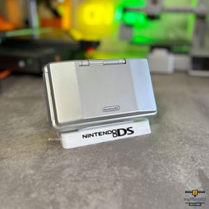 Könnte beinhalten: Eine silberne Nintendo DS Handheld-Spielkonsole auf einem weißen Präsentationsständer. Der Ständer ist auf der Vorderseite mit dem schwarzen Schriftzug NINTENDO DS bedruckt. Die Konsole ist geschlossen und zeigt das Nintendo-Logo auf dem Deckel.